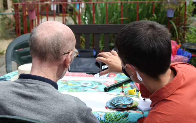 Photo shows two people looking at a mobile phone screen as they take a picture of an artwork they have just made. On the left is an older man with his back to us, he is wearing glasses and a grey top, on the right is a younger man with dark hair, he is holding the phone and is wearing an orange top. In front of them is a table covered with a colourful table cloth and art materials.Essex 2021 Magic Me Workshop 1 Quenby_Photo Credit Georgia Akbar & Lily Ash Sakula